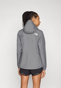 Grijze waterdichte jas met een ritssluiting aan de voorkant, een verstelbare capuchon en elastische manchetten, gedragen over zwarte shorts. Logo is zichtbaar op de achterkant.