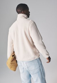 Kremowa kurtka z polaru z wysokim kołnierzem, teksturowaną powierzchnią i długimi rękawami. Połączona z jasnoniebieskimi dżinsami i beżową zamszową torbą.