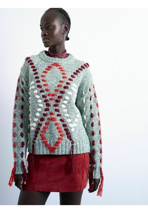 Donna che indossa un maglione grigio testurizzato con motivi intrecciati rossi, bianchi e marrone scuro abbinato a una minigonna in suede rossa, in piedi davanti a uno sfondo bianco.