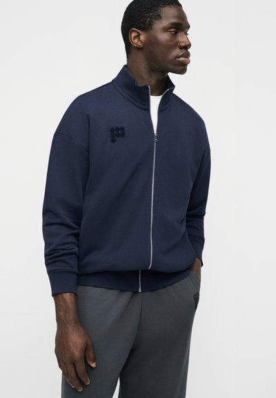 Homme portant une veste zippée bleu marine et un pantalon gris, debout avec une main dans la poche, regardant vers sa droite sur un fond clair uni.