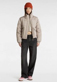 Hellbeige gesteppte Jacke mit hohem Kragen, Frontreißverschluss und Seitentaschen, kombiniert mit schwarzen Jeans mit weitem Bein und pinken Turnschuhen.