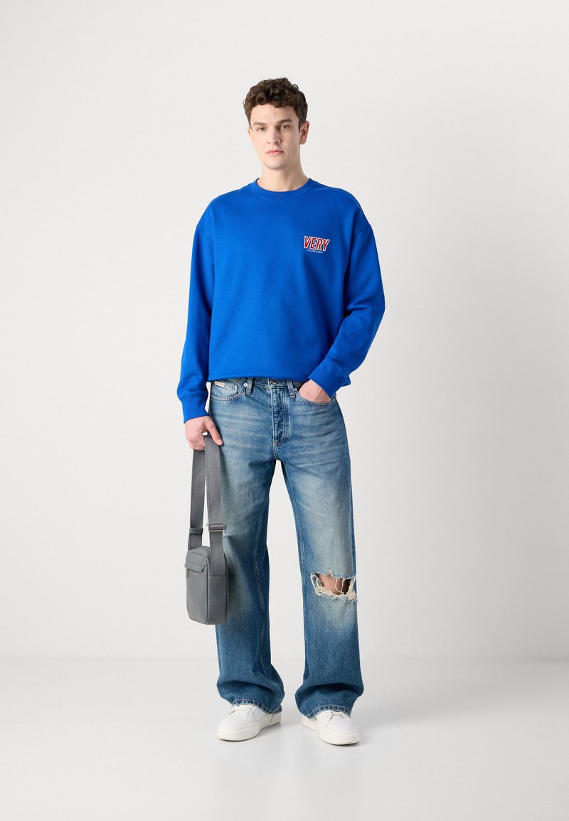 Sudadera de algodón azul con el logo "VERY", combinada con jeans claros desgastados y zapatillas blancas. Una bolsa de hombro gris cuelga a su lado.