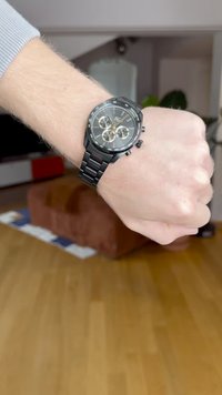 Relógio cronógrafo preto com uma bracelete texturizada preta, mostrador redondo, detalhes em dourado e três submostradores. Usado num pulso em um ambiente de madeira.