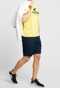 Gul bomullströja med svart text, marinblå shorts, vit jacka över axeln och svarta träningsskor med vita accenter.