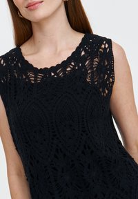 Czarny bezrękawnik z misternym wzorem szydełkowym, falbaniastym dekoltem i teksturowanymi detalami, ukazujący kwiatowe i owalne wzory.