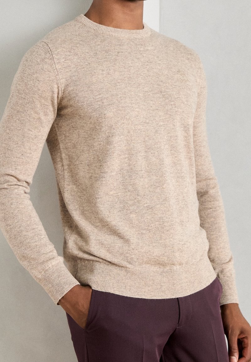 Homme portant un pull beige à col rond et un pantalon violet foncé, une main dans la poche, sur un fond clair uni.