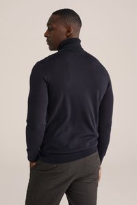 Marinblå polotröja med figurnära passform, ribbad krage och muddar, tillverkad av mjukt tyg, bärs med grå byxor.