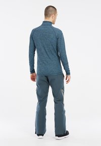 Camicia blu a maniche lunghe con colletto alto, abbinata a pantaloni grigi con zip laterali. Il tessuto ha un aspetto sportivo e texturizzato.
