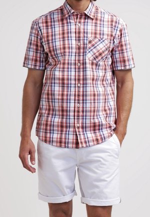 Homme portant une chemise à manches courtes à carreaux rouges et bleus avec des boutons blancs et un short blanc retroussé, debout les mains détendues.