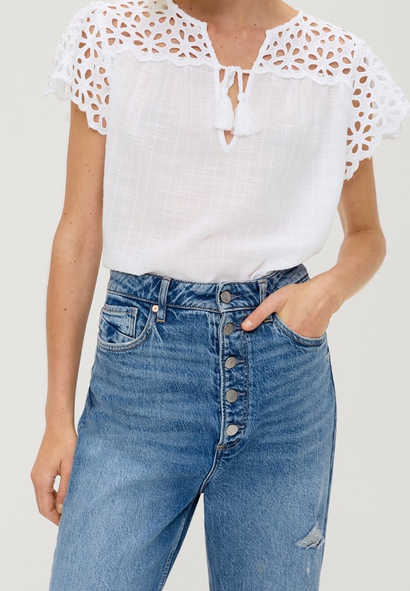 Hvid top med broderede blomstrede søm og nøglehullets detalje, parret med højtaljede blå denim jeans med knaplukning og lomme.