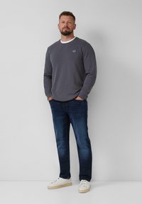 Sweatshirt gris avec des détails de col blanc, jeans bleus et baskets blanches. Le modèle se tient devant un fond uni.
