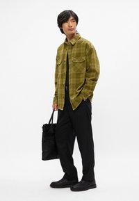 Chemise en flanelle à carreaux verts avec deux poches poitrine, t-shirt noir, pantalon noir ample et chaussures noires ; tenant un sac fourre-tout noir.