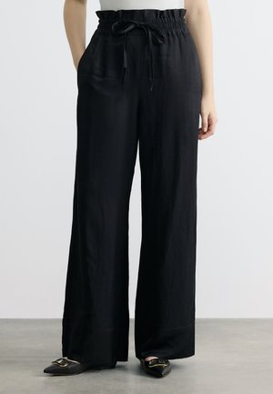 Black high-waisted wide-leg pants with drawstring waist worn with black pointed flats featuring gold buckles.