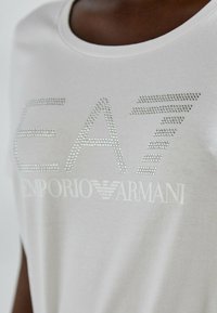 T-shirt grigio chiaro con scollatura rotonda, decorata con il testo "EA7" e "Emporio Armani" in strass, texture in cotone liscia.