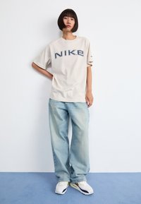 Bež majica s kratkim rukavima marke Nike s velikim plavim logotipom i ležernim krojem, u kombinaciji s svijetloplavim hlačama širokih nogavica i bijelim tenisicama.