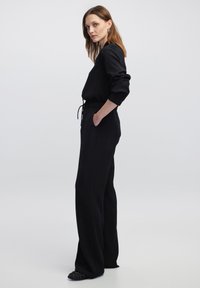 Zwarte jumpsuit met lange mouwen, verstelbare taille, zijzakken en geribbelde manchetten. Beschikt over een strakke, gladde textuur en een relaxte pasvorm.