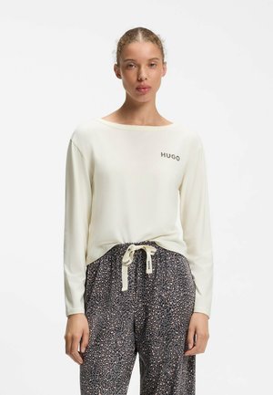 Jonge vrouw draagt een wit shirt met lange mouwen met de tekst "HUGO" en losse broek met luipaardprint en een gestrikte koord taille.
