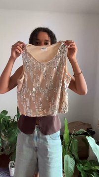 Top sem mangas bege adornado com lantejoulas prateadas, apresentando um decote redondo e um corte solto, segurado contra um fundo neutro com plantas.
