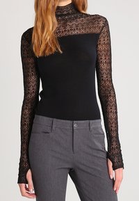 Svart topp med långa, genomskinliga spetsärmar och halvhög krage; kombinerad med grå byxor; detaljerat spetsmönster på ärmarna.