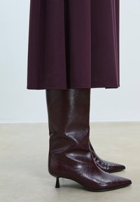Burgundske škornje do kolen z lesketajočo usnjeno obdelavo, s koničastim nosom in ozkim črnim peto, v kombinaciji z ohlapno temno obleko.