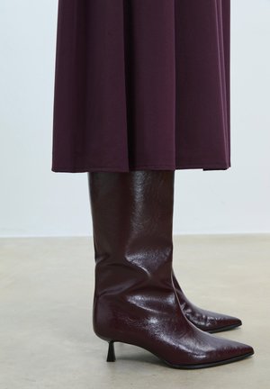 Burgundské kozačky po kolena s lesklým koženým povrchem, špičatou špičkou a štíhlým černým podpatkem, doplněné o splývavé tmavé šaty.