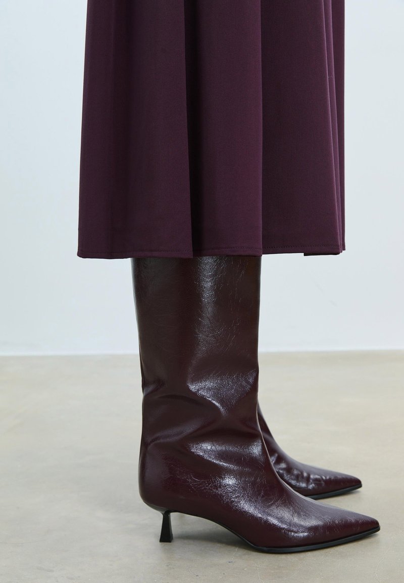 Bottes bordeaux à hauteur de genou avec une finition en cuir brillant, pointe effilée et un talon noir fin, associées à une robe fluide de couleur sombre.
