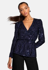 Marineblaue Paillettenbluse mit V-Ausschnitt, langen Ärmeln und gerafften Taillendetails. Sie hat eine strukturierte Oberfläche mit glänzenden, reflektierenden Akzenten.