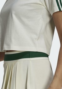 Vit kortärmad t-shirt med gröna detaljer, ihopparad med en plisserad krämfärgad kjol med en grön och vit midjeband. Slät tygtstruktur.