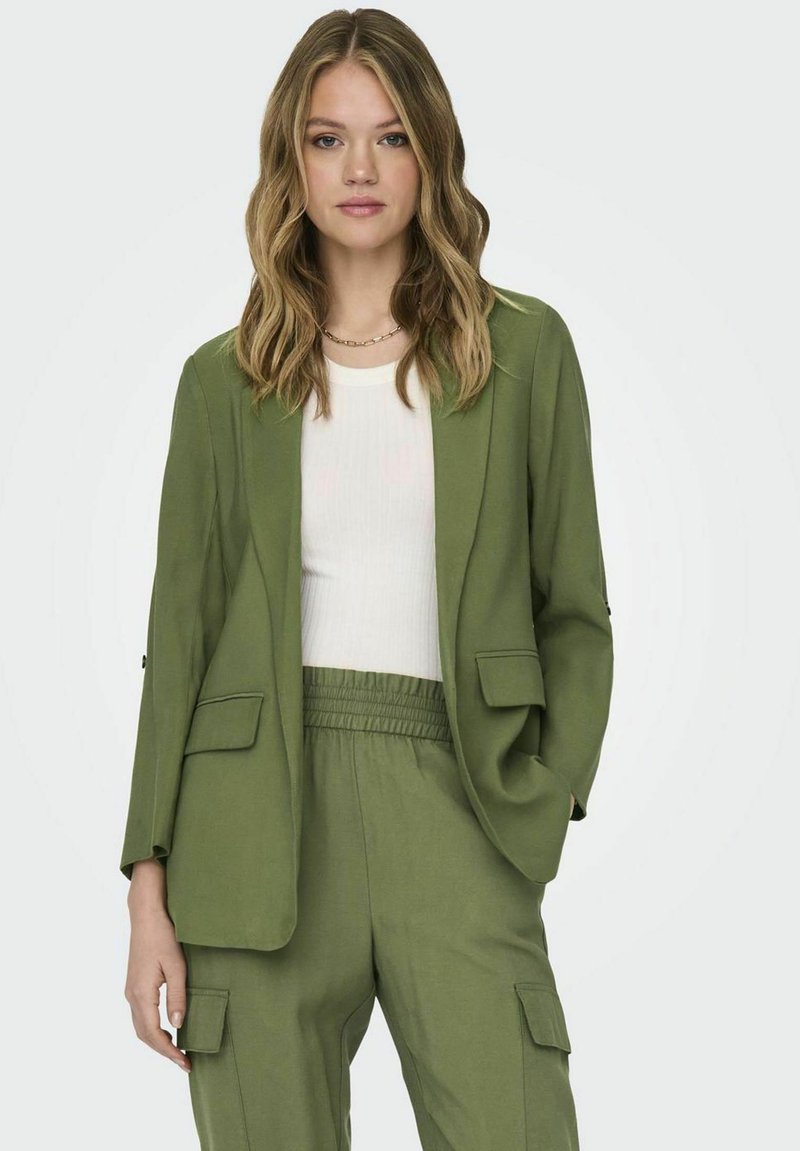 Blazer verde con una vestibilità rilassata, due tasche frontali e tessuto morbido. Indossato su una maglietta bianca a coste, abbinato a pantaloni cargo verdi coordinati.
