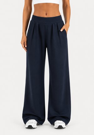 Taille fine portant un pantalon large ample bleu marine foncé avec poches latérales et baskets blanches sur un fond clair uni.