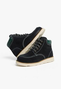 Stivali scamosciati blu scuro con cuciture bianche, punta rotonda e una finitura verde sul tallone. La suola in gomma beige offre trazione.