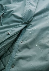 Waterdichte teal jas met een gladde textuur, zichtbare ritsclosure en drukknoopjes, met waterdruppels op het oppervlak.