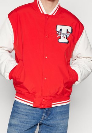 Czerwono-biała kurtka varsity z zatrzaskami, dużą naszywką w kształcie litery "T" i napisem "Tommy" na piersi, noszona z niebieskimi dżinsami i rękami w kieszeniach.