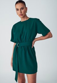 Vihreä lyhythihainen mekko, vyötetty vyötäröltä hopeasella renkaalla. Sujuva kangas, rento istuvuus ja käänteet.