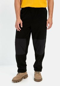 Sort fleecebukser med nylonpaneler i sort, der har en elastisk talje. Båret med beige støvler. Minimalistisk design uden synlige logoer.