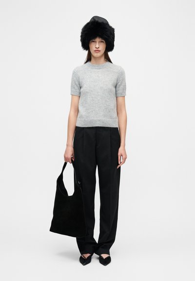 Γυναίκα που φοράει μαύρο γούνινο καπέλο, γκρι πουλόβερ με κοντά μανίκια, μαύρο παντελόνι με φαρδιά μπατζάκια, μαύρα μυτερά παπούτσια και κρατάει μια μεγάλη μαύρη τσάντα tote.