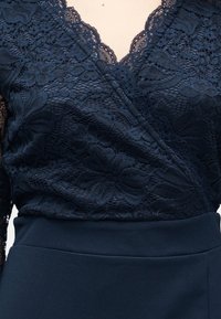 Γυναίκα που φοράει μπλούζα από σκούρο μπλε δαντέλα με λουλουδένιο μοτίβο και σκάλιστο V-λαιμόκοψη, μπλουζωμένη μέσα σε μονοχρωματική σκουρόχρωμη μπλε φούστα.