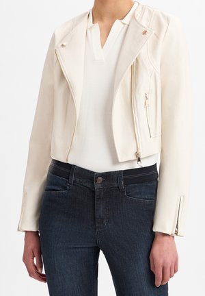 Kvinde iført en hvid læderjakke med lynlås over en hvid skjorte kombineret med mørkeblå jeans mod en ensfarvet baggrund.