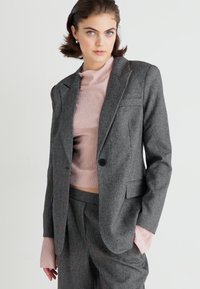 Grijze wollen blazer met notch-revers en een enkele knoopsluiting, gecombineerd met een roze geribde trui die gelaagd gebruik van manchet-details laat zien.