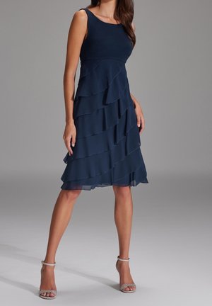 Robe sans manches bleu marine avec volants superposés, corsage ajusté et ourlet à hauteur du genou. Tissu lisse avec des accents transparents en bas.