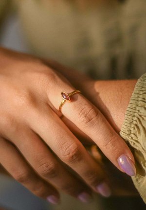 Anello d'oro con una gemma viola a forma di marquise su un sottile band, esposto su una mano con pelle chiara e unghie morbide e arrotondate.