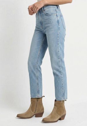 Person mit hellblauen, hoch sitzenden Jeans und hellbraunen Wildleder-Stiefeletten mit Holzabsätzen, stehend vor einem schlichten weißen Hintergrund.