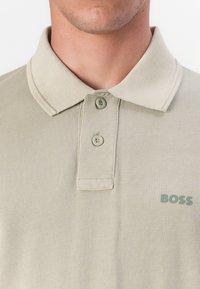 Beige poloshirt med teksturert stoff, to knapper ved kragen, og en liten grønn "BOSS" logo på høyre side.