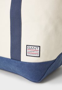 Sac en toile crème avec des accents bleu foncé. Il présente une étiquette GANT, une forme rectangulaire, une construction robuste et des coutures visibles.
