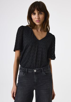 Schwarzes Oberteil mit V-Ausschnitt, kurzen Ärmeln und strukturiertem Zickzackmuster. Kombiniert mit dunkelgrauen Jeans, die eine taillierte Silhouette zeigen.
