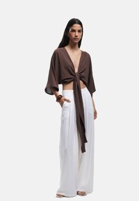 Kvindelig iført en brun dyb V-hals binde-front top med brede ærmer, hvide brede bukser, brun armbånd og hvide sandaler, stående mod en hvid baggrund.