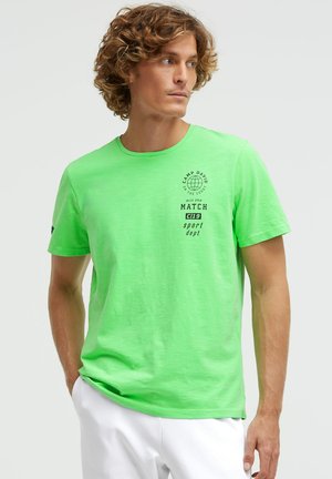 Man with curly hair wearing bright green Camp David graphic t-shirt and white pants, looking sideways with one hand in pocket.