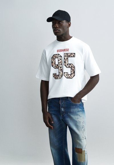 Homme portant un t-shirt blanc surdimensionné avec l'impression léopard "95" et le texte rouge "DSQUARED2", jean bleu déchiré avec des patchs, et casquette noire.