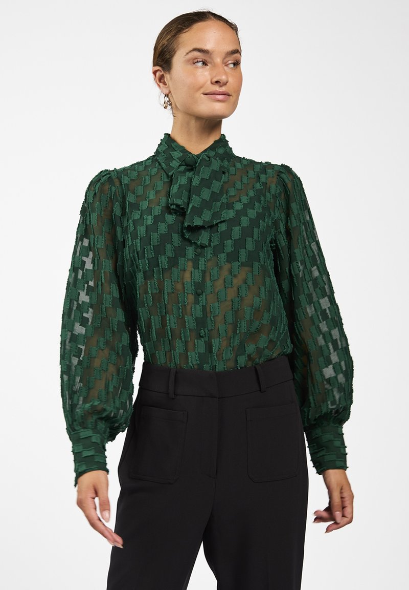 Groene doorzichtige blouse met textuurpatroon, hoge hals en strik. Heeft pofmouwen en een knoopsluiting, gecombineerd met zwarte getailleerde broeken.