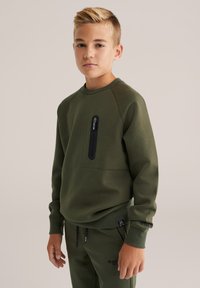 Olívazöld pulóver, kerek nyakkivágással, raglán ujjakra és elülső cipzáras zsebbel. Puha tapintású és laza szabású. Része egy összeillő szettnek.